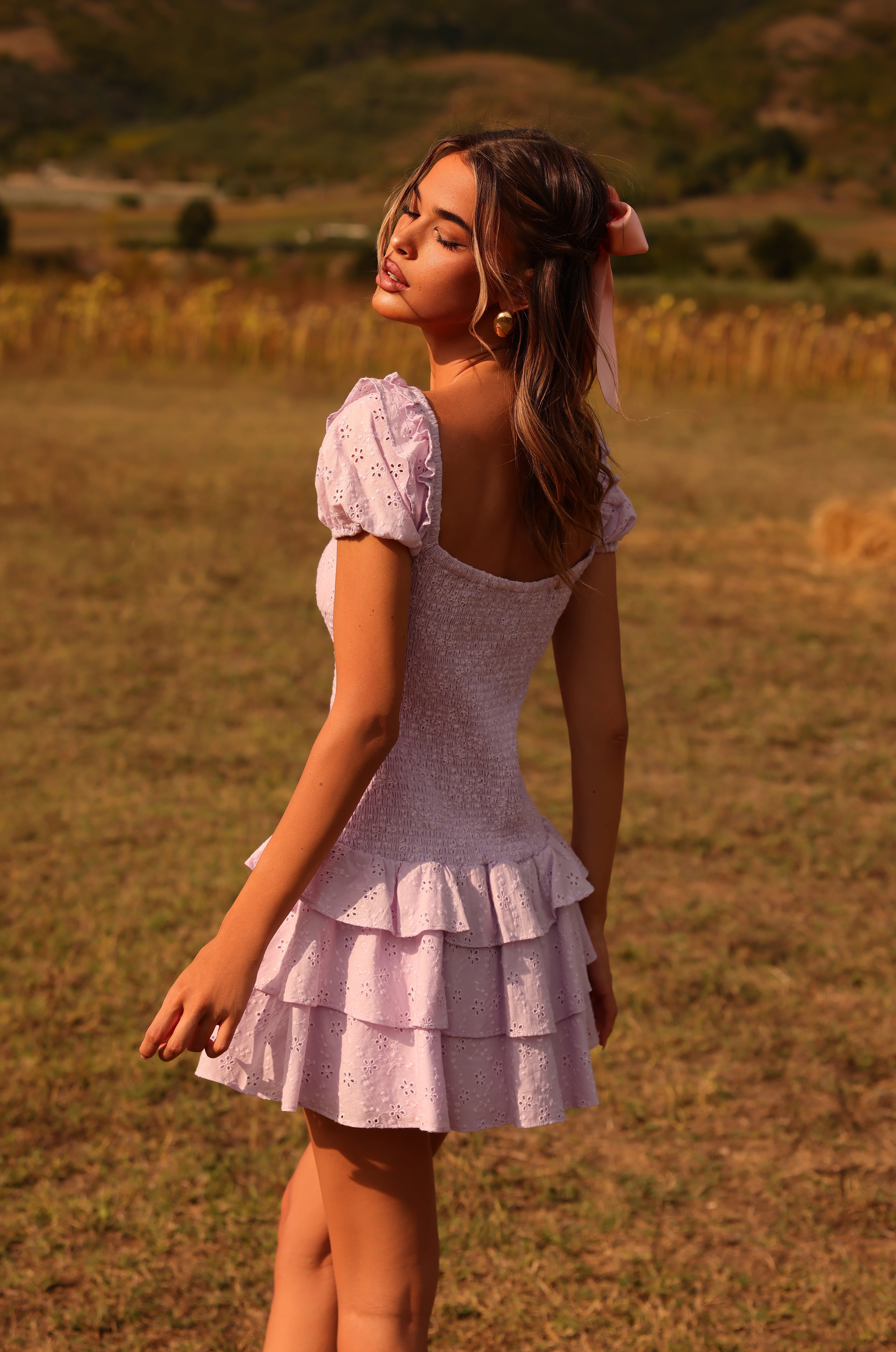 A woman in the Lilac Purple Short Summer Dress: Macie, featuring ruffled details, stands on a grassy field outdoors, looking over her shoulder with a neutral expression.